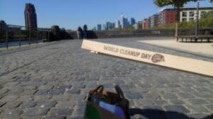 Ein Holzstab mit der Aufschrift „World Cleanup Day“ wird vor dem gepflasterten Mainufer und der Frankfurter Skyline in die Kamera gehalten.