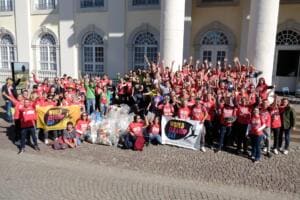 Eine große Gruppe in roten Shirts jubelt mit World-Cleanup-Day-Bannern und vielen gefüllten Säcken vor einem Gebäude mit Säulen und Rundbogenfenstern.