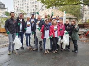 Eine Gruppe von Erwachsenen in World-Cleanup-Day-Shirts und grünen Handschuhen steht mit weißen Säcken und Greifzangen auf einem nassen Hof.