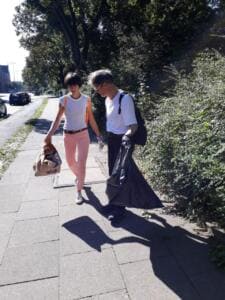 Eine Frau und ein Mann in weißen Shirts gehen mit vollen Säcken einen Gehweg entlang, neben ihnen eine Hecke.