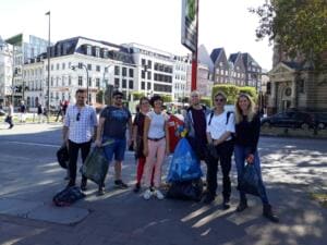 Eine Gruppe von Erwachsenen steht mit vollen Säcken an einer Straßenecke vor Stadthäusern mit Backsteingiebeln.