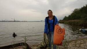 Eine Frau in blauer Jacke steht am Ufer eines breiten Gewässers und hält ein großes orangefarbenes Fundstück und ein Tau, im Hintergrund Hafenkräne.