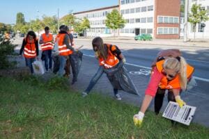 Mehrere Erwachsene in orangefarbenen Warnwesten sammeln mit Säcken Müll auf einem Grünstreifen neben einer Straße vor Bürogebäuden.