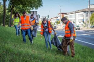 Fünf Helfer in Warnwesten sammeln mit schwarzen Säcken Müll auf einer Grasböschung an einer Hauptstraße.