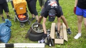 Helfer sortieren auf einer Wiese gesammelte Glasflaschen, Dosen und einen alten Reifen in Kartons und Säcke.