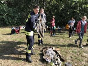 Ein junger Feuerwehrmann zieht ein großes verdrecktes Stück Plastikfolie aus dem Gebüsch, im Hintergrund Kinder auf einer Wiese.