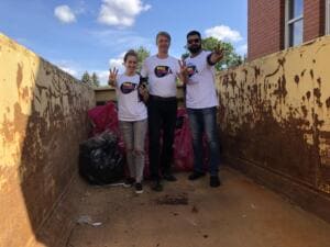 Drei Erwachsene in World-Cleanup-Day-Shirts stehen gut gelaunt in einem großen rostigen Container neben vollen Säcken und zeigen das Victory-Zeichen.
