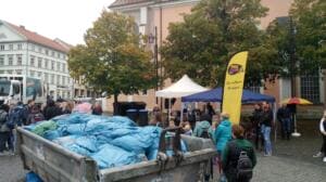 Ein großer Container ist randvoll mit blauen Müllsäcken auf dem Marktplatz, daneben viele Menschen, Pavillons und eine World-Cleanup-Day-Beachflag.