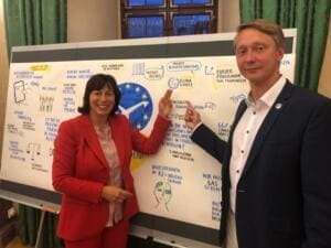 MdEP Marion Walsmann im roten Anzug und Holger Holland zeigen gemeinsam auf eine gezeichnete Themenwand, auf der auch der World Cleanup Day steht.