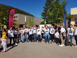 Eine Gruppe in World-Cleanup-Day-Shirts steht zwischen zwei Beachflags auf einem Platz vor modernen Gebäuden und bewaldeten Hängen.