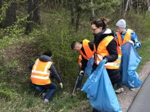 Mehrere Helfer in Warnwesten, darunter ein Kind, sammeln mit blauen Säcken im Graben neben einer Straße.
