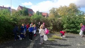 Eine Gruppe Kinder und einige Erwachsene sammeln mit weißen Beuteln Müll auf einem Platz vor Büschen und Häusern.
