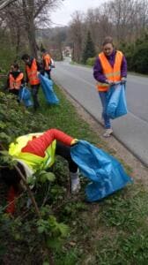 Mehrere Helfer in Warnwesten sammeln mit blauen Säcken Müll an einer Böschung entlang einer Landstraße am Ortsrand.