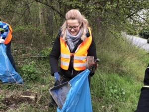 Eine lächelnde Frau in Warnweste hält eine gefundene Metallschale und einen blauen Sack an einer bewachsenen Böschung.