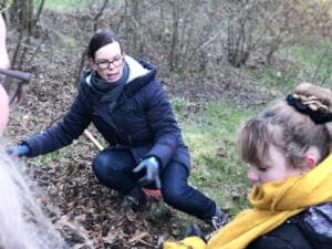 Eine Frau mit Brille und Schal sammelt in einem Waldstück Müll aus dem Laub, vorn ein Mädchen mit gelbem Schal.