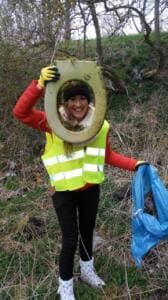Eine lachende Frau in Warnweste hält einen gefundenen Toilettensitz vor ihr Gesicht wie einen Bilderrahmen, in der anderen Hand einen blauen Sack.