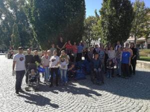 Eine große Gruppe aus Erwachsenen, Kindern und einem Rollstuhlfahrer steht auf einem gepflasterten Platz vor Bäumen und vielen blauen Säcken.