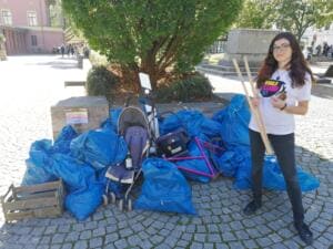 Eine junge Frau im World-Cleanup-Day-Shirt steht mit Holzstäben neben einem Berg aus blauen Säcken, einem Kinderwagen und einem Fahrradrahmen.