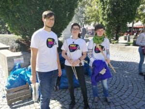Ein Mann und zwei Frauen in World-Cleanup-Day-Shirts stehen mit Holzstäben vor vollen blauen Säcken auf einem Platz.