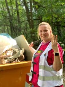 Eine lächelnde Frau in World-Cleanup-Day-Weste zeigt drei Finger vor einem orangefarbenen Container voller Müll im Wald.