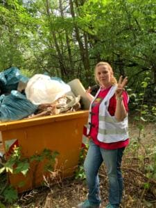 Eine Frau in World-Cleanup-Day-Weste steht im Wald neben einem orangefarbenen Container, der mit Säcken und Sperrmüll gefüllt ist, und zeigt drei Finger.