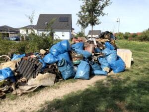 Ein großer Haufen aus blauen Müllsäcken, Wellplatten, Bauschutt und Holz liegt auf einer Wiese vor Einfamilienhäusern.