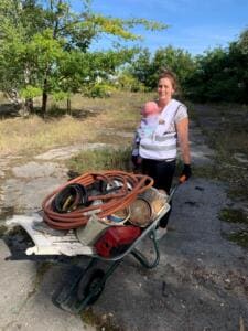 Eine Frau in World-Cleanup-Day-Weste trägt ein Baby in der Trage und schiebt eine Schubkarre voller Gartenschläuche, Metallteile und Sperrmüll über einen alten Betonweg.