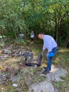 Ein Mann in weißem Hemd hebt im Wald zwischen Bauschutt einen alten Koffer auf, im Hintergrund sammeln weitere Personen.
