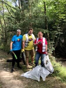 Drei Erwachsene in World-Cleanup-Day-Shirts und Westen stehen mit Greifzangen und vollen weißen Säcken an einem Waldweg.