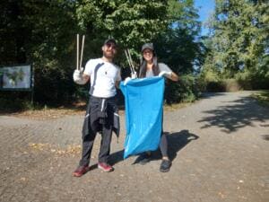 Ein Mann und eine Frau mit Kappen halten Greifzangen hoch und einen blauen Müllsack auf einem gepflasterten Parkweg.