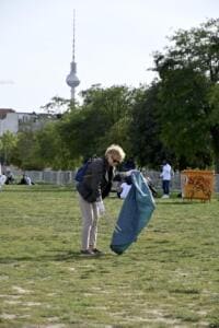 Eine Frau sammelt mit Greifzange und blauem Sack Müll auf einer Wiese im Mauerpark, im Hintergrund der Berliner Fernsehturm.