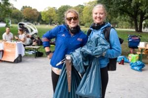 Zwei lachende junge Frauen mit Kopfhörern halten volle blaue Müllsäcke und eine Greifzange, im Hintergrund ein Infostand im Park.