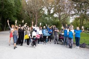 Eine große Gruppe mit Masken reckt jubelnd die Arme in die Höhe, viele halten Säcke und Schilder, im Hintergrund Bäume des Parks.