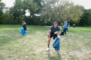 Ein junger Mann mit Kopfhörern hebt auf einer großen Wiese in der Hasenheide Müll auf und hält einen blauen Sack, im Hintergrund weitere Sammelnde.