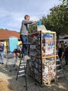 Ein Mann steht auf einer Leiter und füllt einen großen Gitterkasten, der mit gesammeltem Plastikmüll gefüllt ist, daneben Menschen und ein Einkaufswagen.