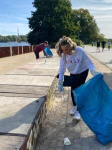 Eine junge Frau in weißem Pullover sammelt mit Greifzange und blauem Sack Müll an einer Uferpromenade, im Hintergrund Wasser und ein Fernsehturm.