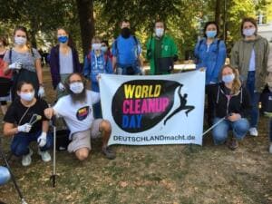 Eine Gruppe mit Masken steht und kniet mit Greifzangen hinter einem World-Cleanup-Day-Banner im Park.