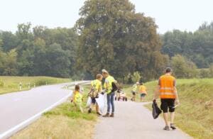 Eine Gruppe in gelben und orangefarbenen Warnwesten sammelt Müll entlang eines Feldwegs neben einer Landstraße, im Hintergrund Bäume.
