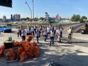 Eine Gruppe in weißen Warnwesten steht auf einer Uferstraße vor Industrieanlagen und Silos, vorne liegen viele orangefarbene Müllsäcke.