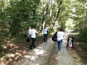 Erwachsene und ein kleines Kind mit Kinderwagen gehen von hinten mit Säcken und Eimern einen sonnigen Waldweg entlang.