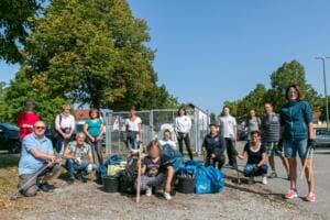 Eine Gruppe aus Erwachsenen und einem Kind sitzt und steht mit Eimern, Greifzangen und blauen Säcken auf einem Parkplatz.