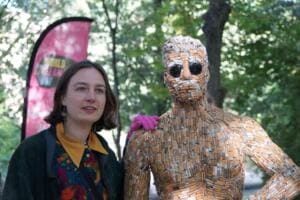 Eine junge Frau steht neben der Skulptur aus Zigarettenstummeln und legt ihr eine Hand auf die Schulter, im Hintergrund eine pinke Beachflag.