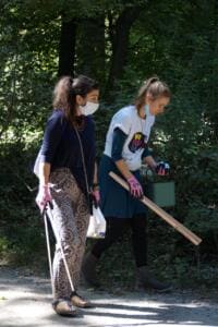 Zwei Frauen mit Masken, Greifzangen und Eimer sammeln Müll an einem schattigen Parkweg.
