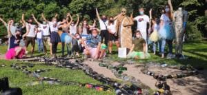 Eine große Gruppe mit Masken jubelt hinter einem Bild aus gesammelten Glasflaschen auf der Wiese, daneben die Skulptur aus Zigarettenstummeln und eine Figur aus Dosen.