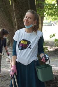 Eine junge Frau im World-Cleanup-Day-Shirt mit Maske am Kinn hält einen grünen Eimer und eine Greifzange im Park.