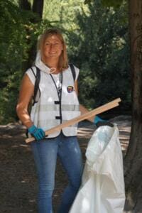 Eine lächelnde Frau in World-Cleanup-Day-Weste hält eine Holzgreifzange und einen weißen Sack unter Bäumen.