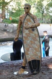 Eine lebensgroße Figur aus gesammelten Zigarettenstummeln mit Sonnenbrille und Surfbrett steht am Eisbach, im Hintergrund Surfer auf der Welle.