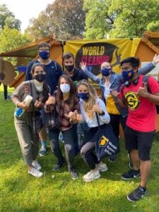 Eine Gruppe junger Leute mit Masken posiert fröhlich auf einer Wiese vor einem World-Cleanup-Day-Banner, eine Person trägt eine Tasche der Munich Business School.