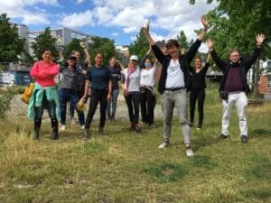 Eine Gruppe Erwachsener mit Masken reckt jubelnd die Arme auf einer Wiese, im Hintergrund ein Bürogebäude.