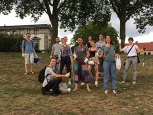 Eine Gruppe Erwachsener mit Greifzangen und Säcken steht unter Bäumen auf einer Wiese vor historischen Gebäuden.